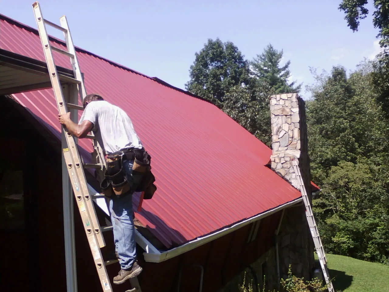 Roofer on a ladder beside a red metal roof and stone chimney — Soffit & Fascia Repair by Roofing Brothers in Port Angeles